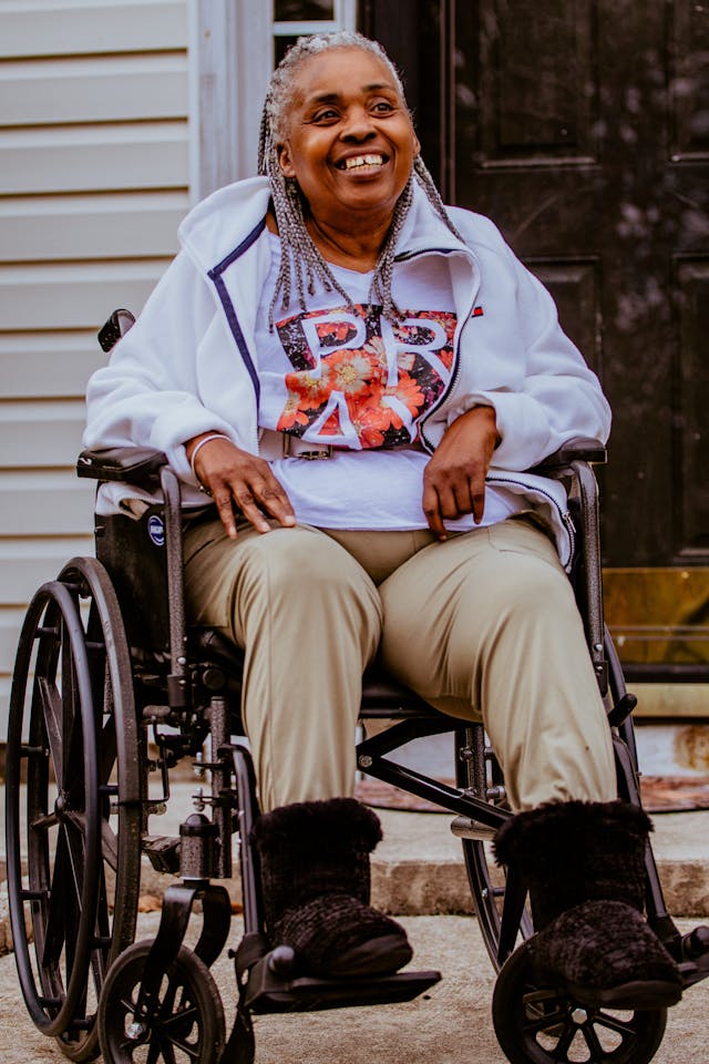 Smiling elderly woman sitting in a wheelchair outdoors, wearing a white jacket and graphic t-shirt